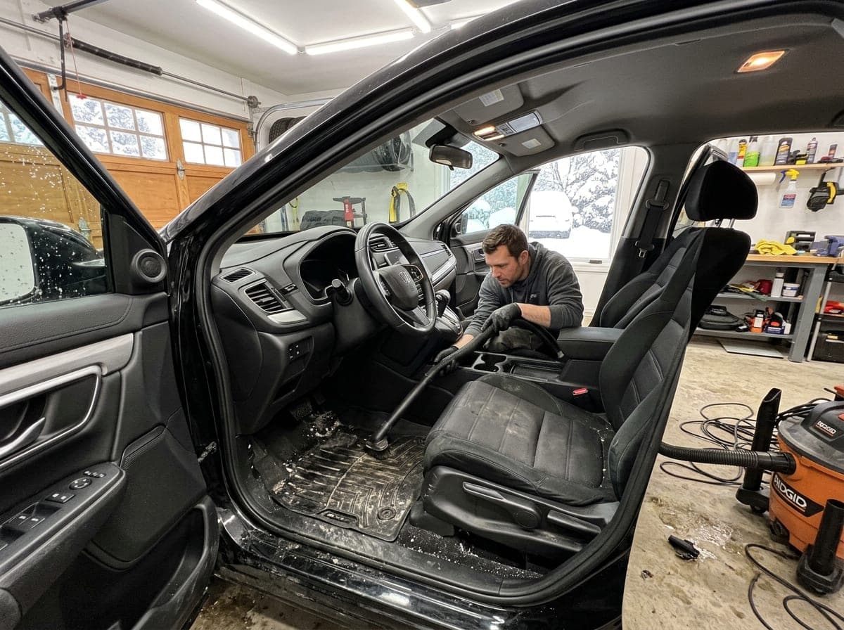 A detailer cleans the interior of a car with winter mats in a bright heated garage, with snow visible through the window.