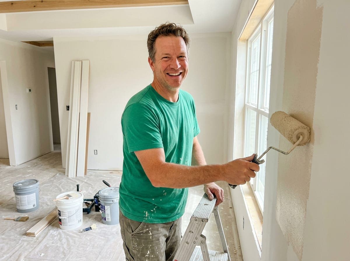 Painter rolling white paint onto a wall from a step ladder in a bright room.