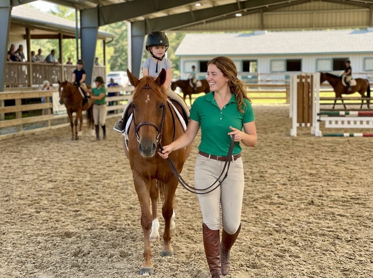 An instructor in a riding helmet helps a young rider on a horse along a fenced arena, with a barn and hay bales in the background.